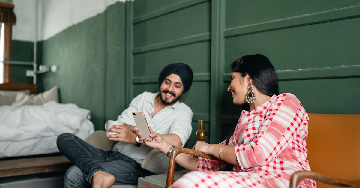 Positive smiling Indian couple in casual traditional outfit relaxing on chairs and browsing mobile phone together in green bedroom