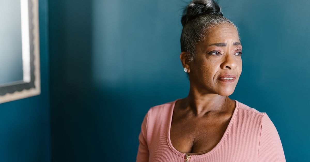 Elderly woman in pink gazing thoughtfully by a blue wall indoors.
