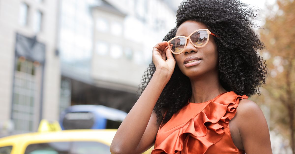 Fashionable woman poses outdoors in urban setting, wearing a striking orange dress and sunglasses.