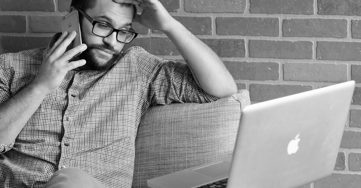 A man in glasses looking worried while on a phone call, sitting by a laptop in an office.