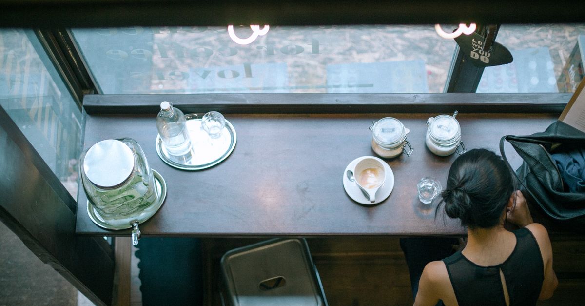 From above back view of faceless woman enjoying cup of hot coffee while spending time in modern cafeteria in daytime