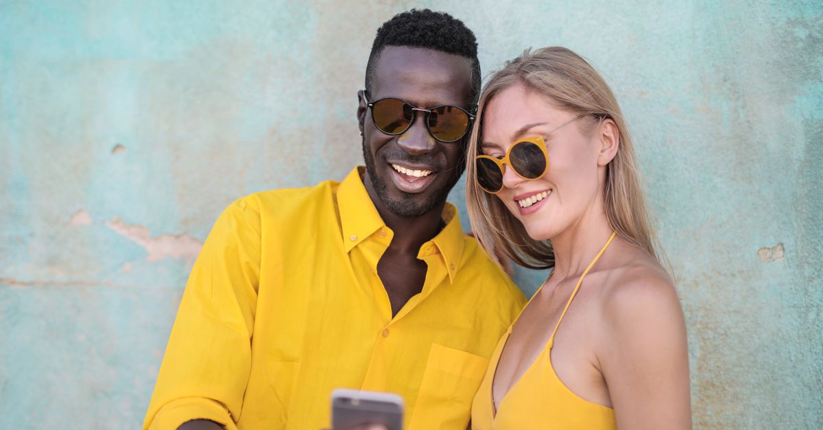 Smiling couple in bright yellow outfits taking a selfie against a textured turquoise wall.